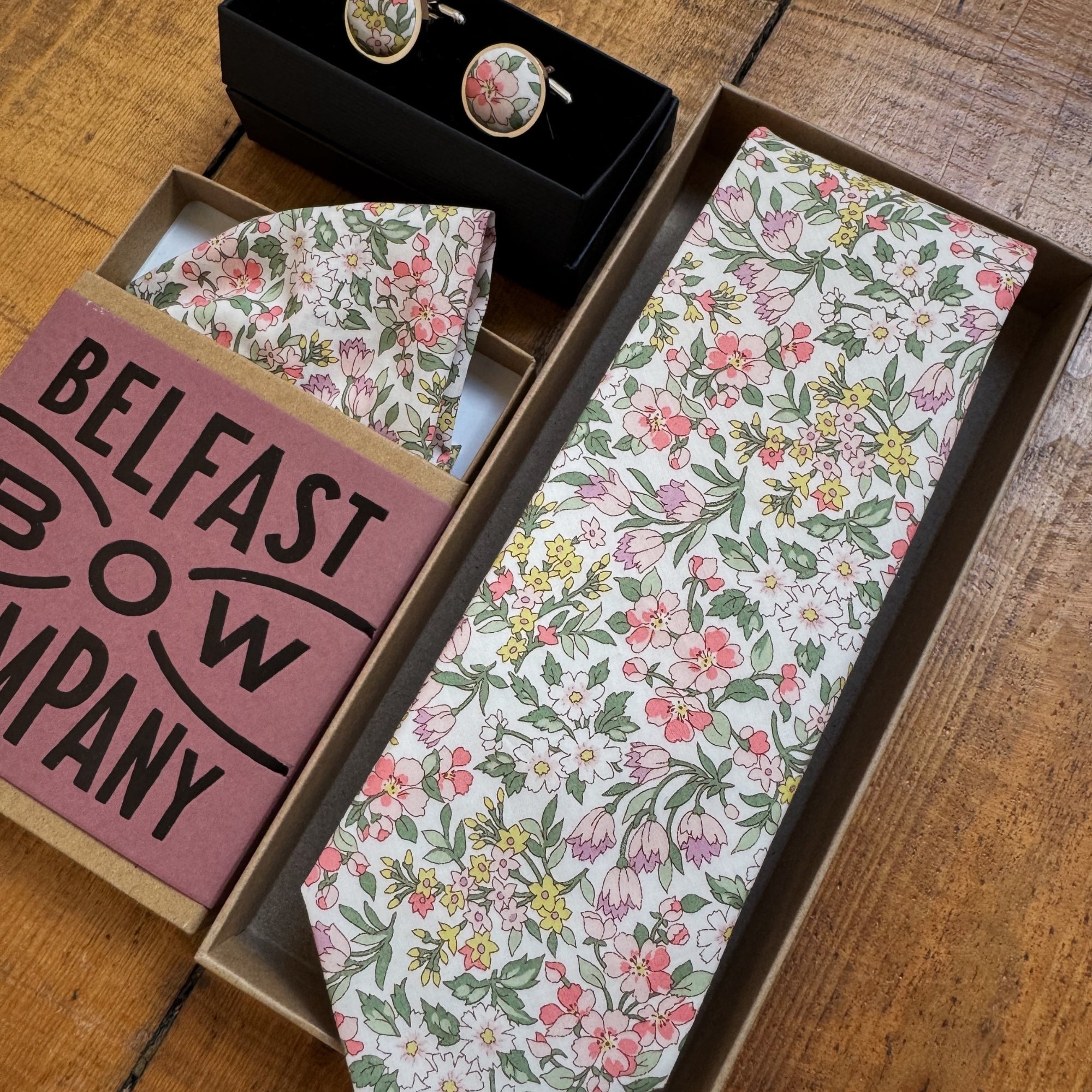 Floral patterned tie and cufflinks set with 'Belfast Bow Company' branding on a wooden surface.