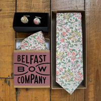 Floral tie, pocket square, and cufflinks in a box labeled 'Belfast Bow Company' on a wooden surface.
