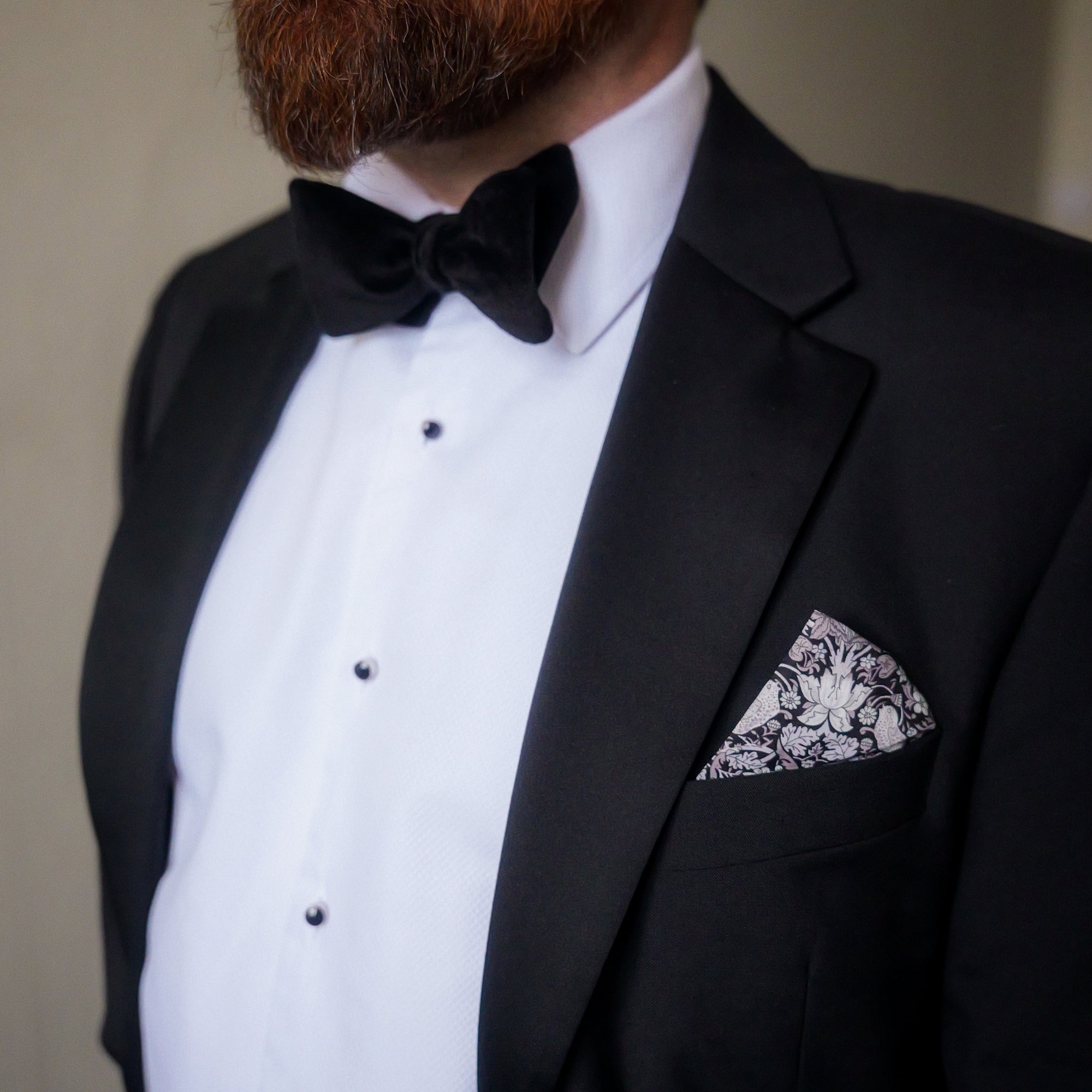 Man wearing a black tuxedo with a white shirt and bow tie, featuring a decorative pocket square.