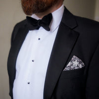 Man wearing a black tuxedo with a white shirt and bow tie, featuring a decorative pocket square.